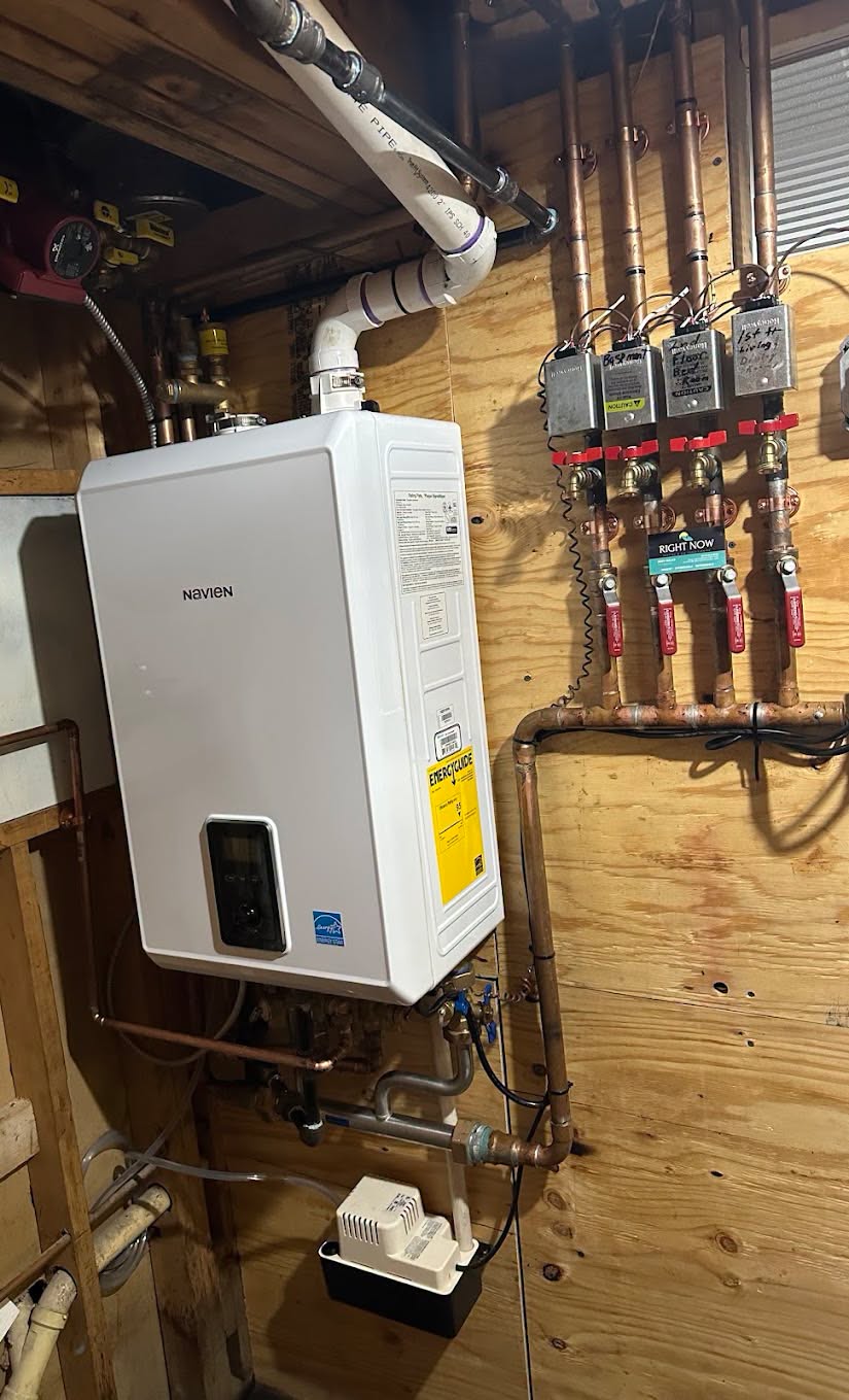 A white Navien tankless water heater is mounted on a wooden wall with copper pipes and multiple valves connected.