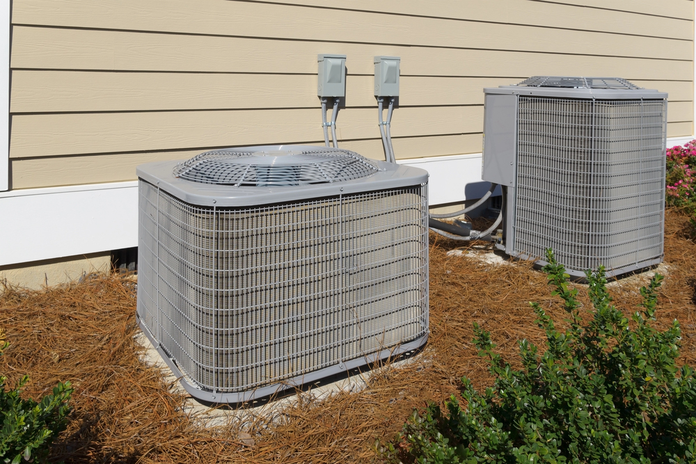 Two outdoor air conditioning units are installed on concrete pads next to a beige building, surrounded by mulch and green shrubs.