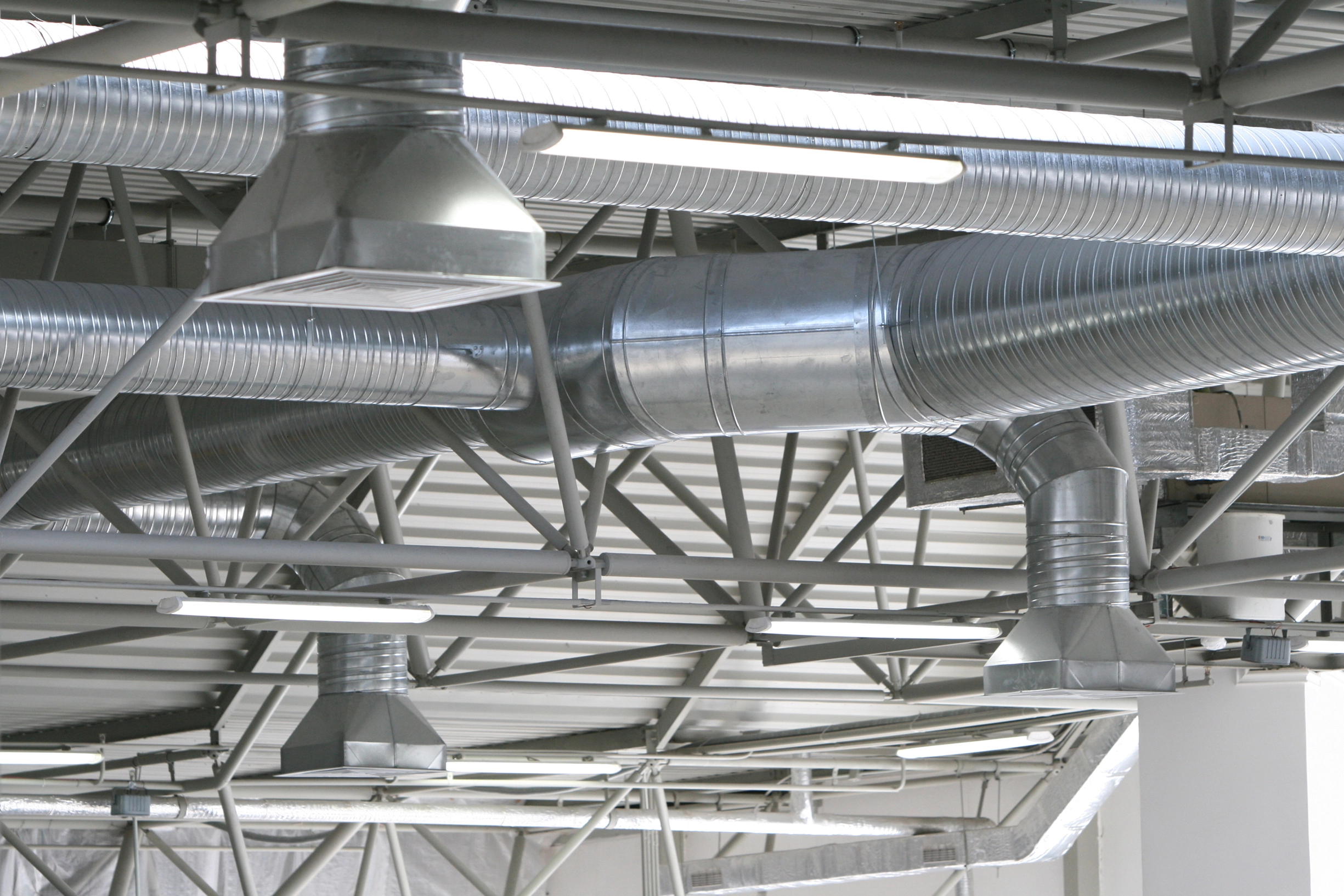 Shiny metal air ducts and vents are visible on the ceiling of an industrial or commercial building, with exposed beams and light fixtures.