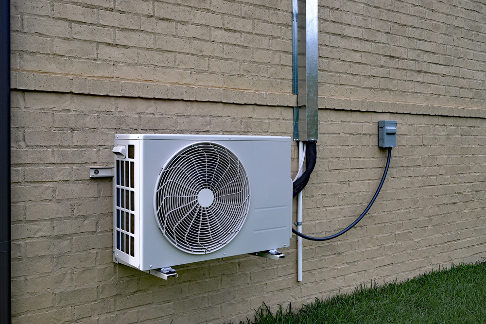 A white outdoor air conditioning unit is mounted on a beige brick wall, with electrical cables running to a nearby utility box above a grass lawn.