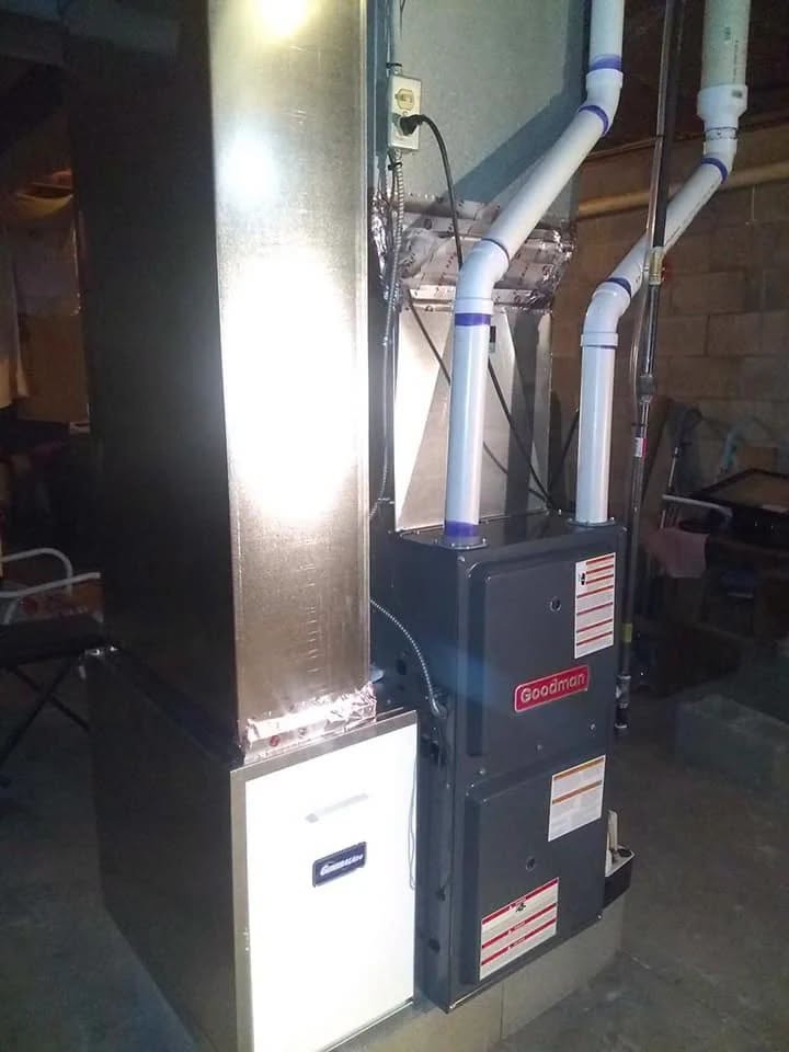 A Goodman furnace unit installed in a basement with connected ductwork and PVC pipes, positioned next to a Carrier-branded air handler.