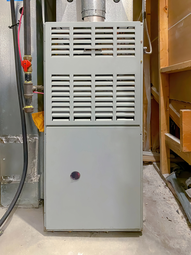 A gray furnace unit with vented panels installed in a basement, surrounded by pipes and unfinished wooden framing.