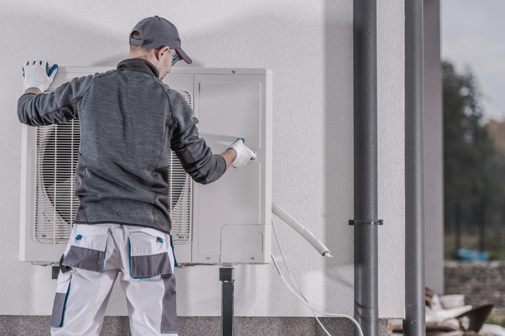 A technician wearing a cap and gloves is servicing or inspecting an outdoor air conditioning unit mounted on a wall.