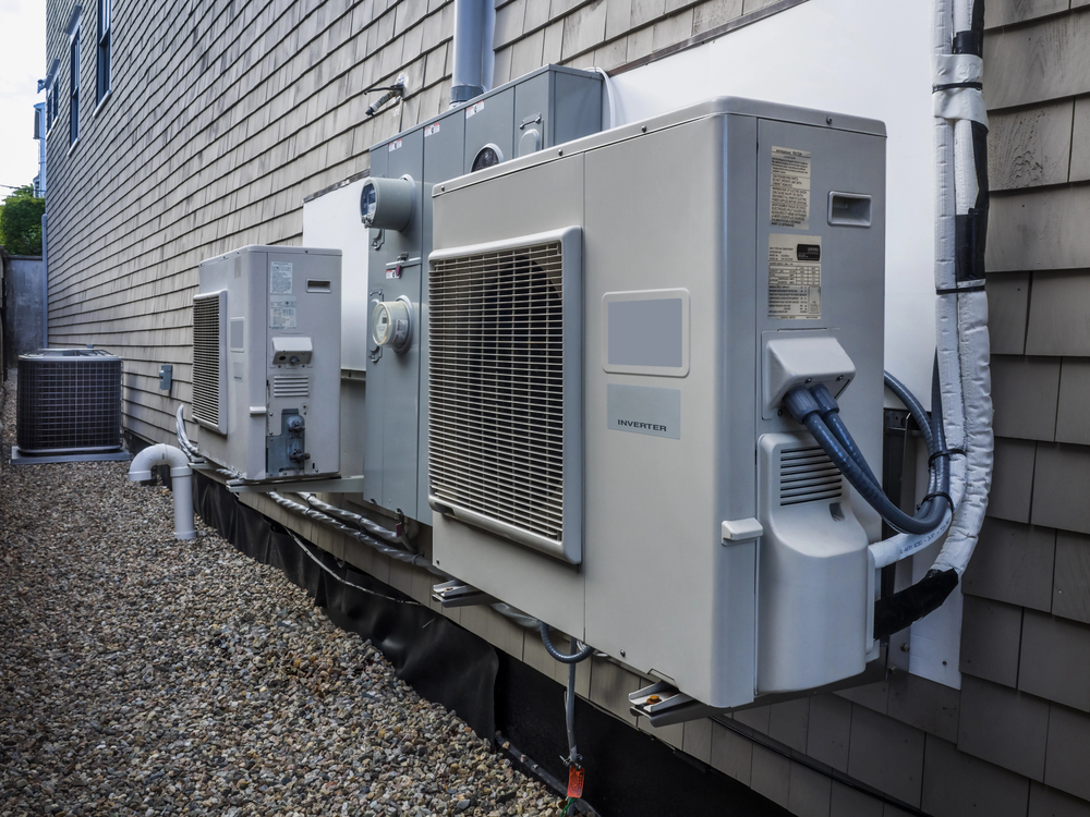 Several outdoor HVAC units are mounted on a building’s exterior wall above a gravel-covered ground.
